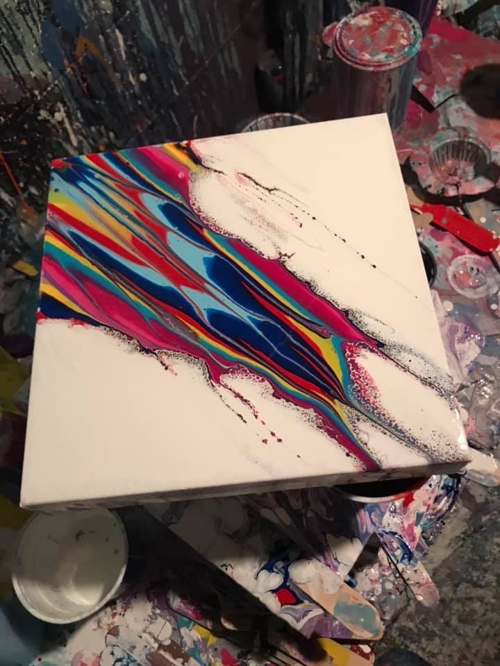 A white canvas with a diagonal sweep of magenta, pink, blue, and black paint, sitting on a paint-splattered studio table.